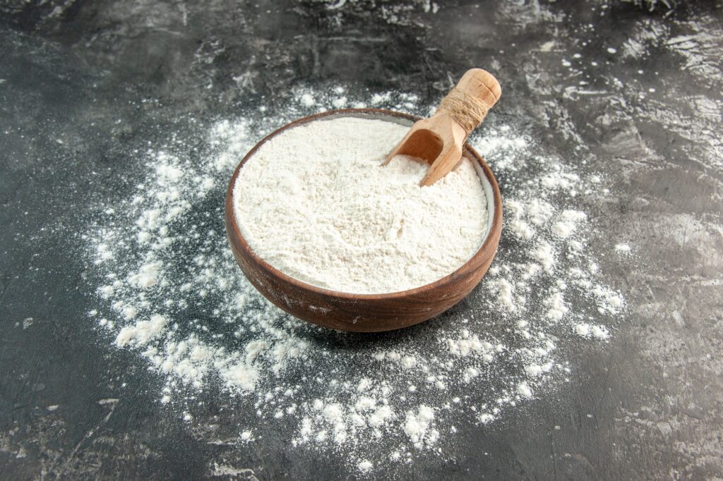 Front close view of white flour with wooden spoon inside and outside of brown bowl on gray
